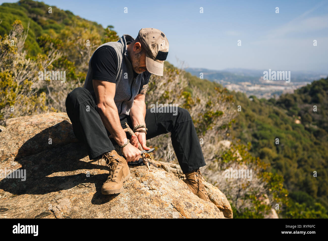 Man sitting on a rock in the nature with great view carving a stock of a tree with a knife Stock Photo