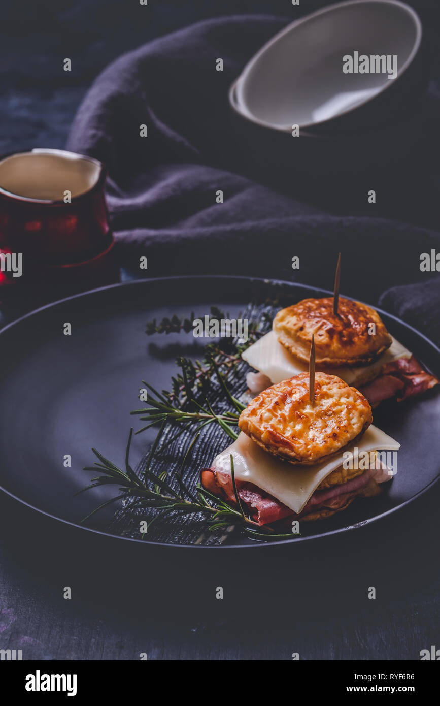 Vertical photo with two sandwiches. Sandwiches are from puff pastry dough squares stuffed by cheese and dried ham. Food is placed on black plate with  Stock Photo