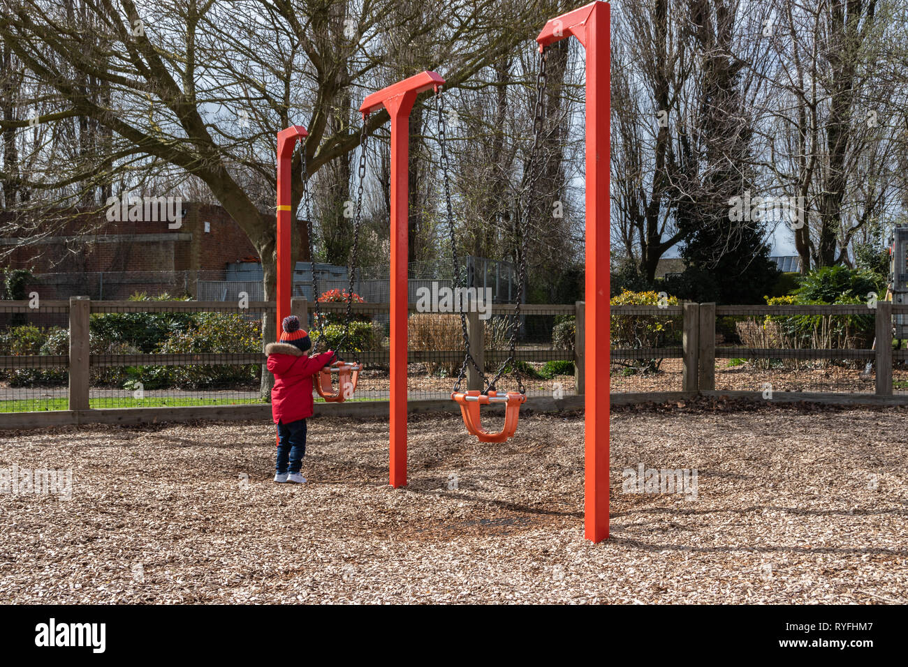 Two year old boy playing with swings in the park wearing a bobble hat and red coat Stock Photo