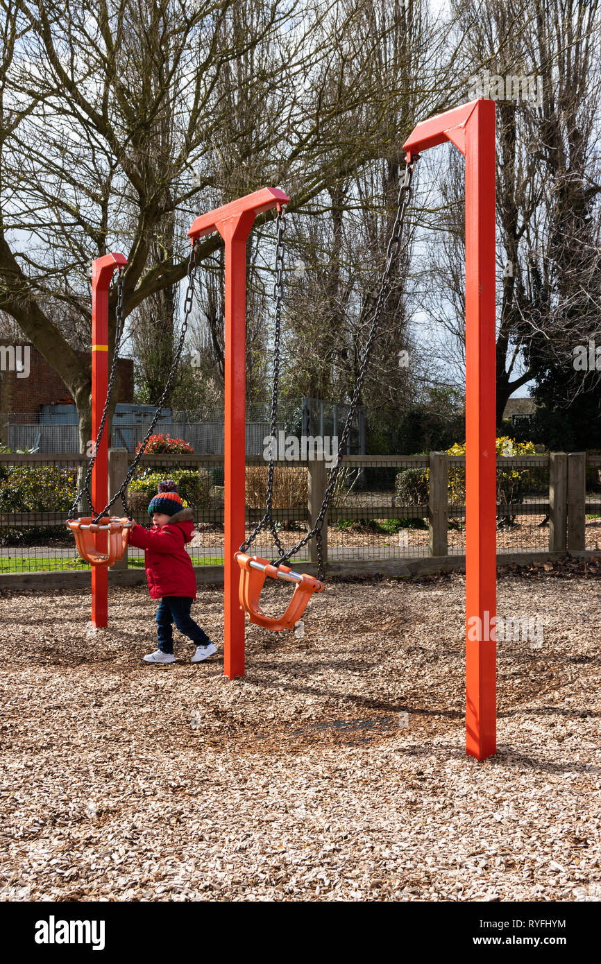 Two year old boy playing with swings in the park wearing a bobble hat and red coat Stock Photo