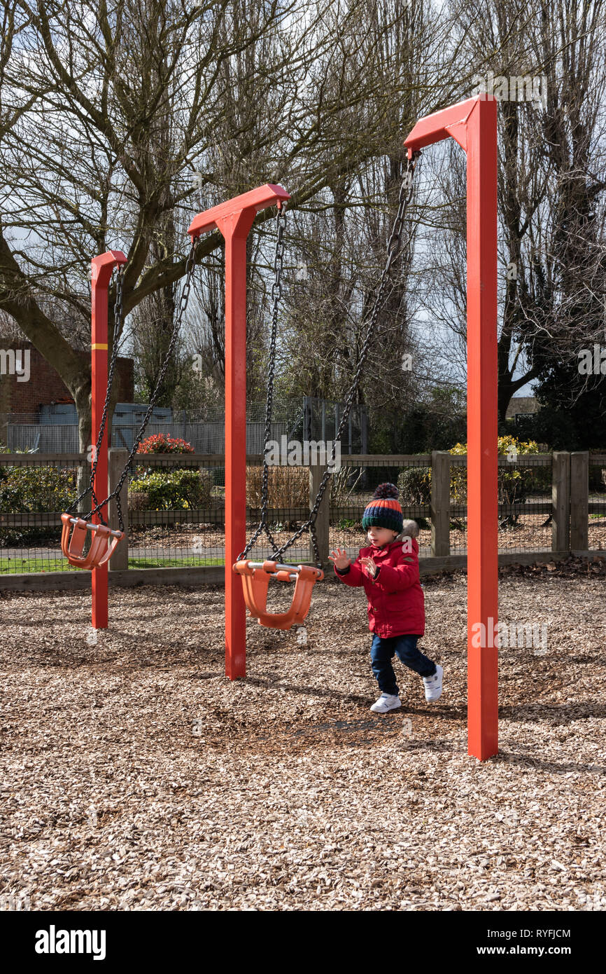 Two year old boy playing with swings in the park wearing a bobble hat and red coat Stock Photo