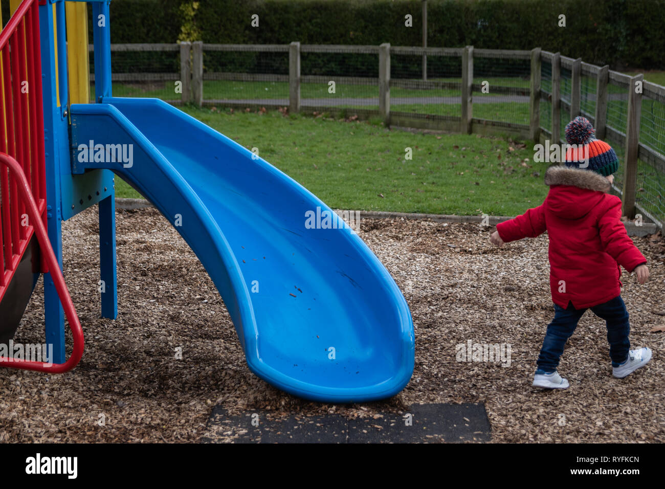 Two year old boy playing on the plastic slide at the park wearing a red coat and bobble hat Stock Photo