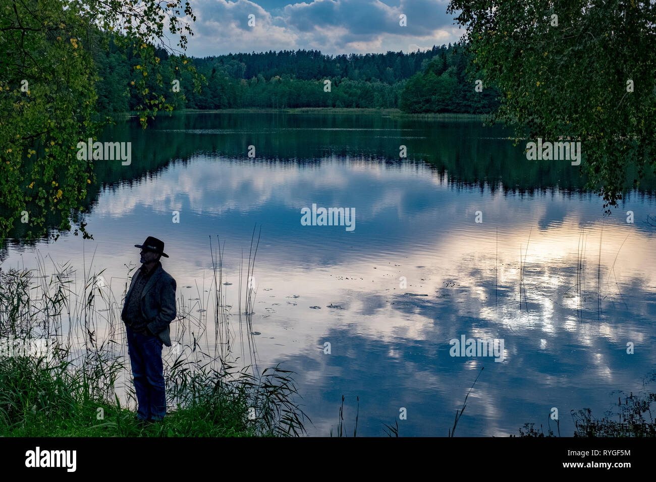 Man in hat is silhouetted as he stands by the side of the water near sunset in the lake district of northeastern Poland near Suwalki Stock Photo