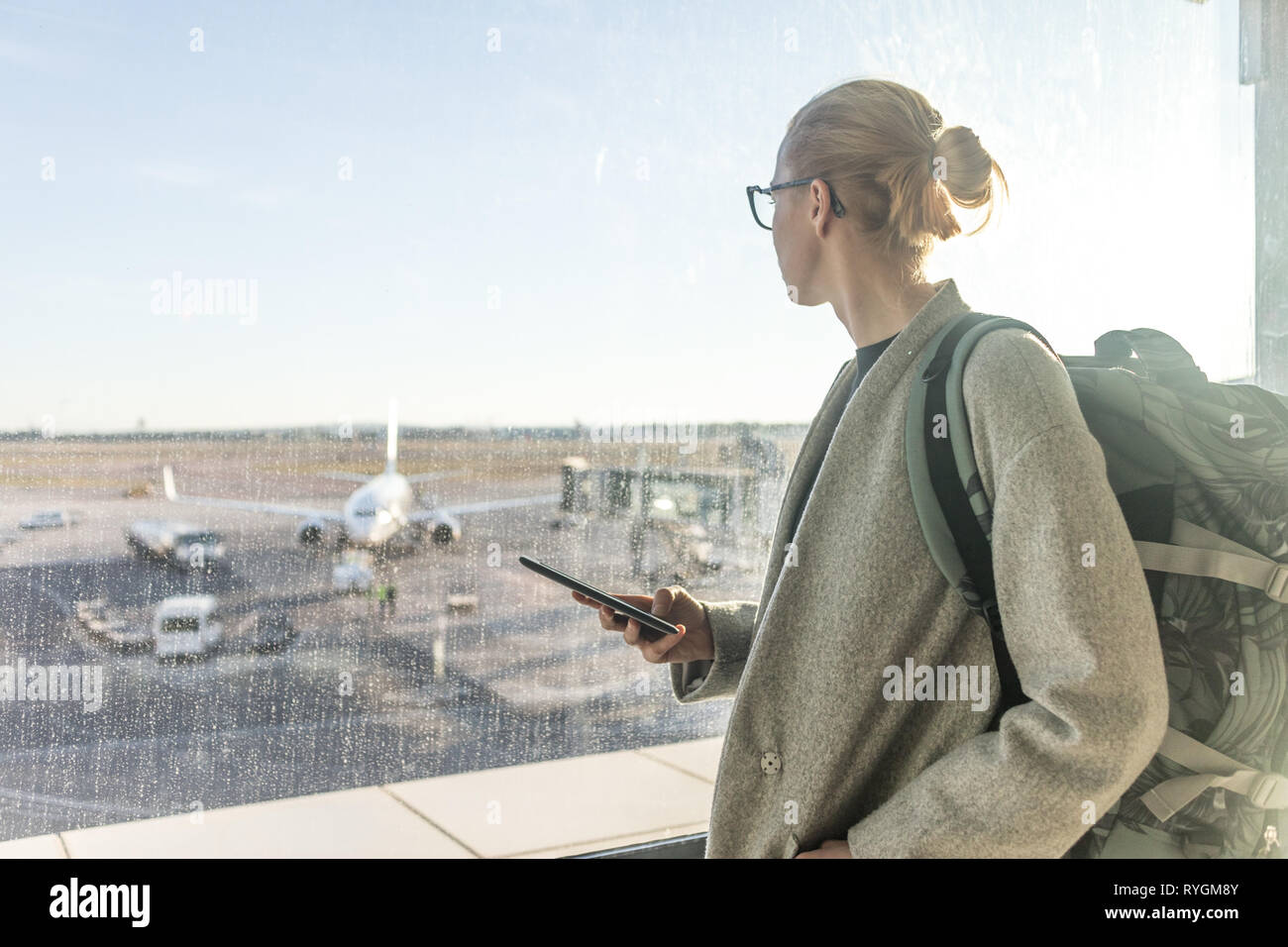 Casually dressed female traveler at airport, holding smart phone device, looking through the airport gate windows at planes on airport runway Stock Photo