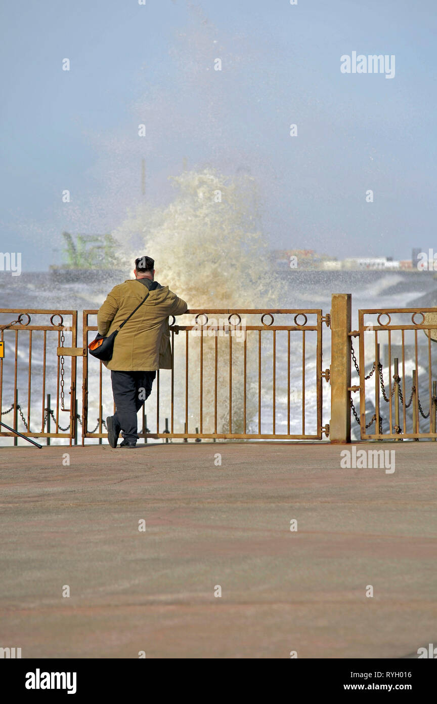 Man leaning against metal gate watching waves break on to the seawall,Blackpool,UK Stock Photo
