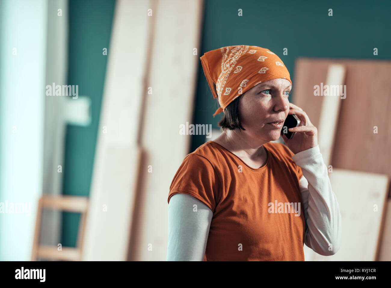 Female carpenter with head kerchief during serious mobile phone conversation with client in carpentry woodwork workshop Stock Photo