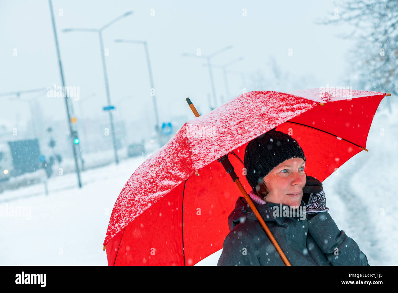 Contented woman talking on mobile phone under red umbrella while standing in the street in winter snow Stock Photo