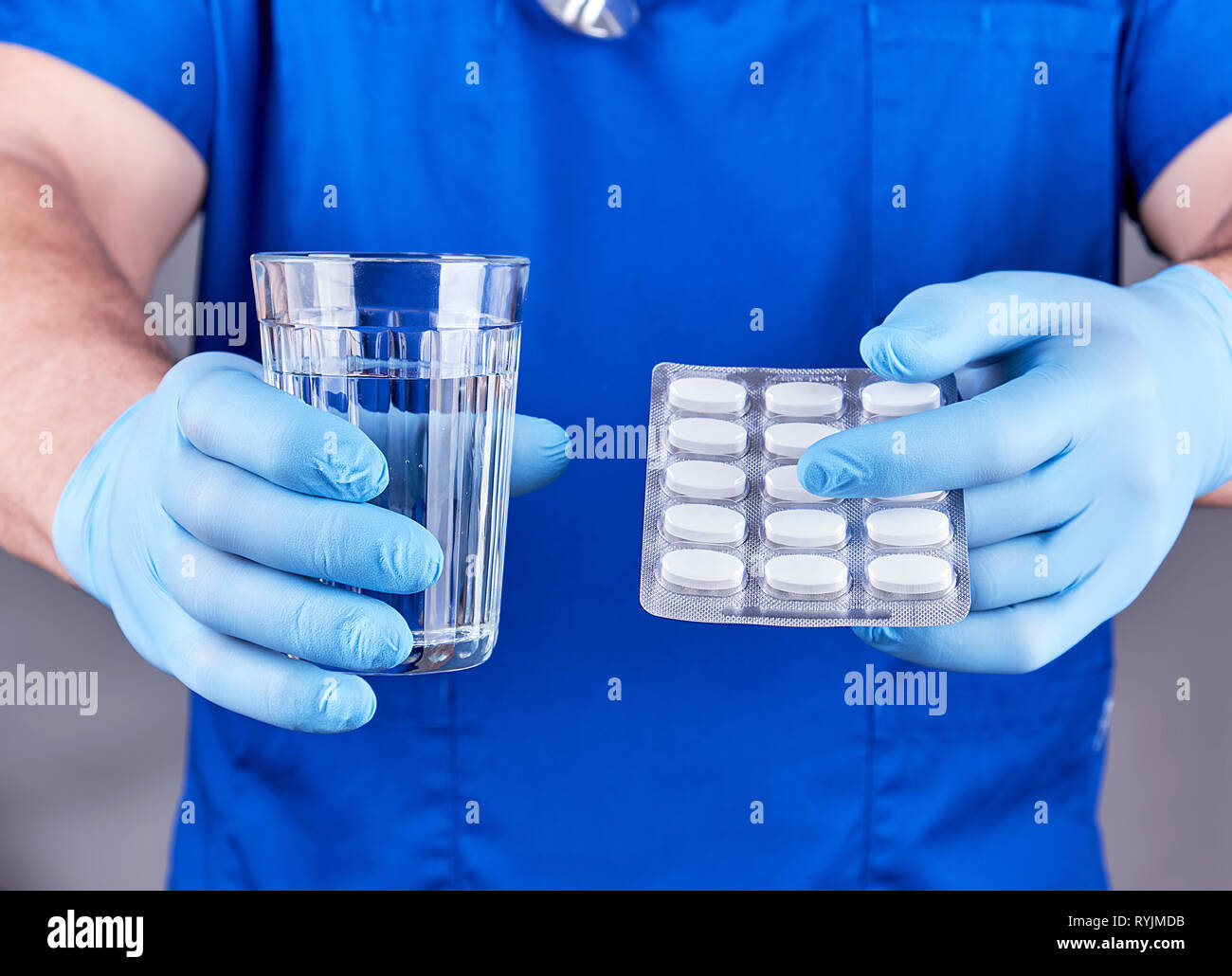 doctor in blue uniform and sterile latex gloves holds a glass of water and a pack of white pills Stock Photo