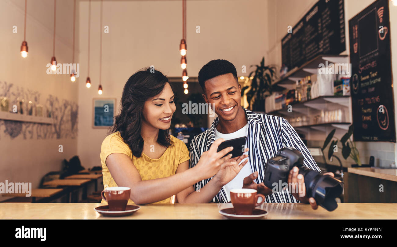 Young woman taking pictures from male friends digital camera. Young man and woman sitting at coffee shop with smart phone and dslr camera. Stock Photo