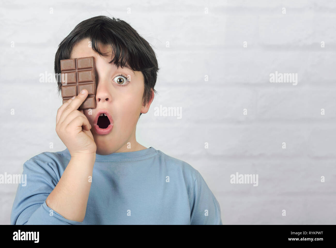 surprised child who covers an eye with an chocolate bar against brick background Stock Photo