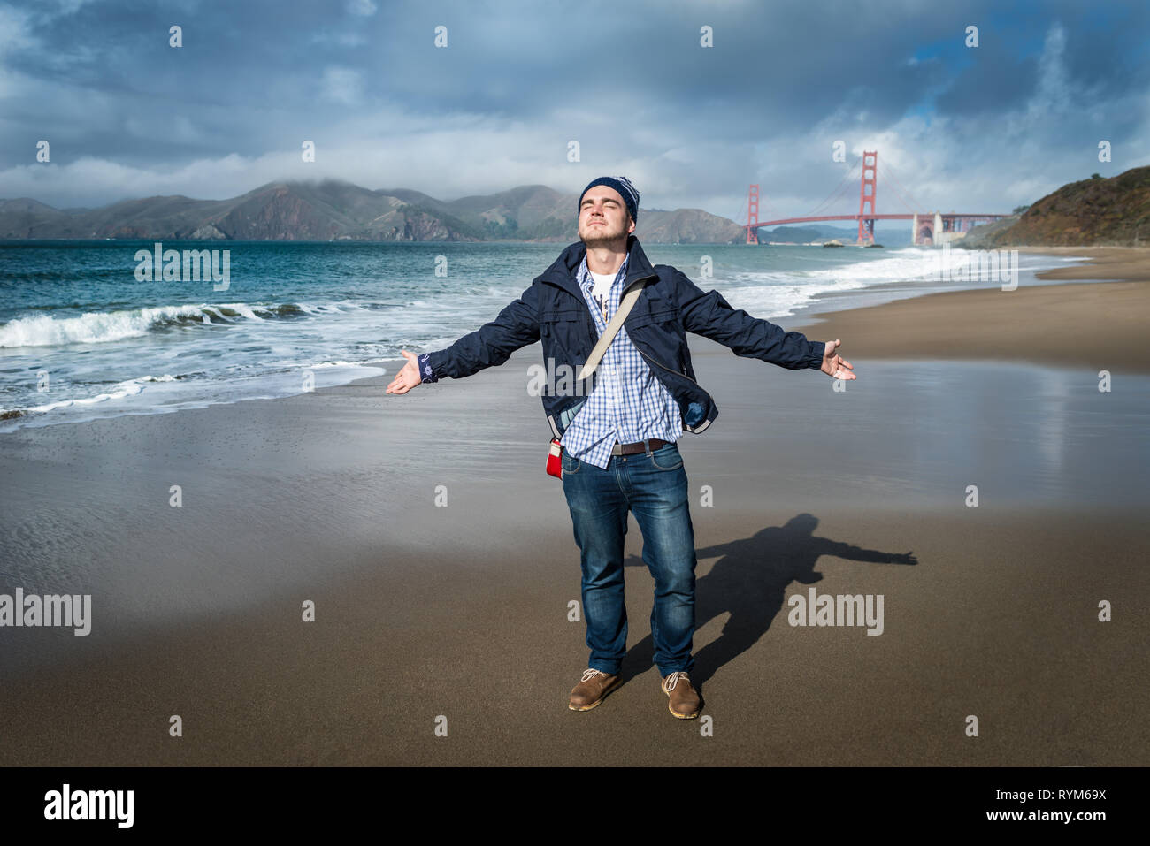 Young man standing on the beach with view on famous bridge in San Francisco, California. Handsome guy gesticulating and enjoying holidays. Stock Photo