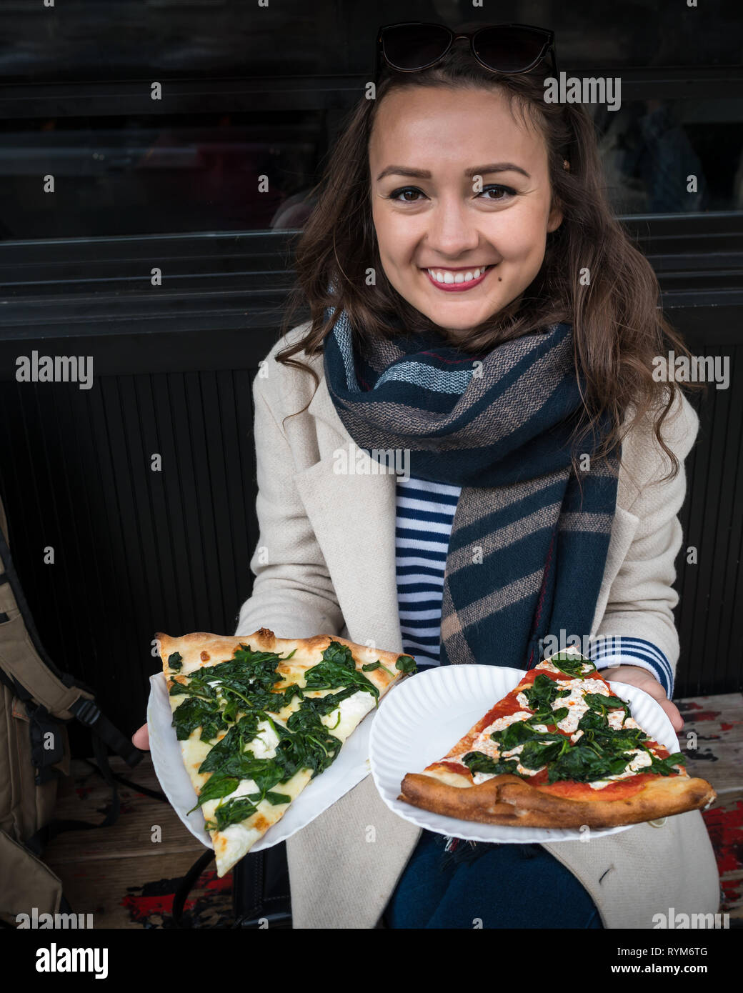 Happy young woman looking at camera, smiling and holds two big pieces of juicy pizza. Elegant girl sitting outdoor and offer to try pizza in New York Stock Photo
