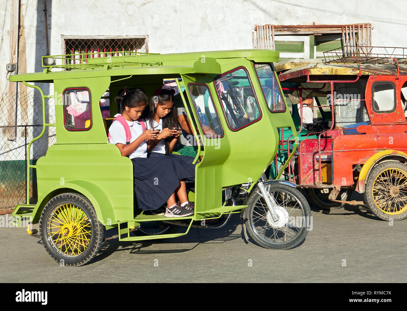 Romblon, Romblon Province, Philippines: Colorful tricycle in front of an old house with two student girls using their mobile phone Stock Photo