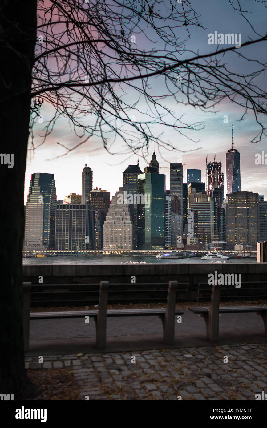 Scenic view from Brooklyn on Manhattan skyscrapers in evening time in New York, USA. Benches on riverside on background of city downtown and sunset. Stock Photo