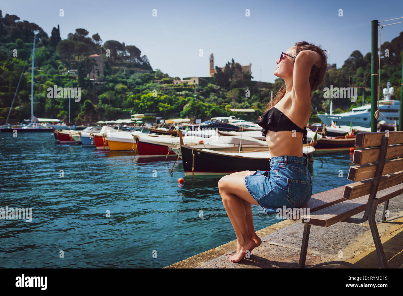 Young barefoot woman sits on bench in marina in Portofino, Italy. Female tourist enjoying sunny day on background of small village, sea and boats. Stock Photo