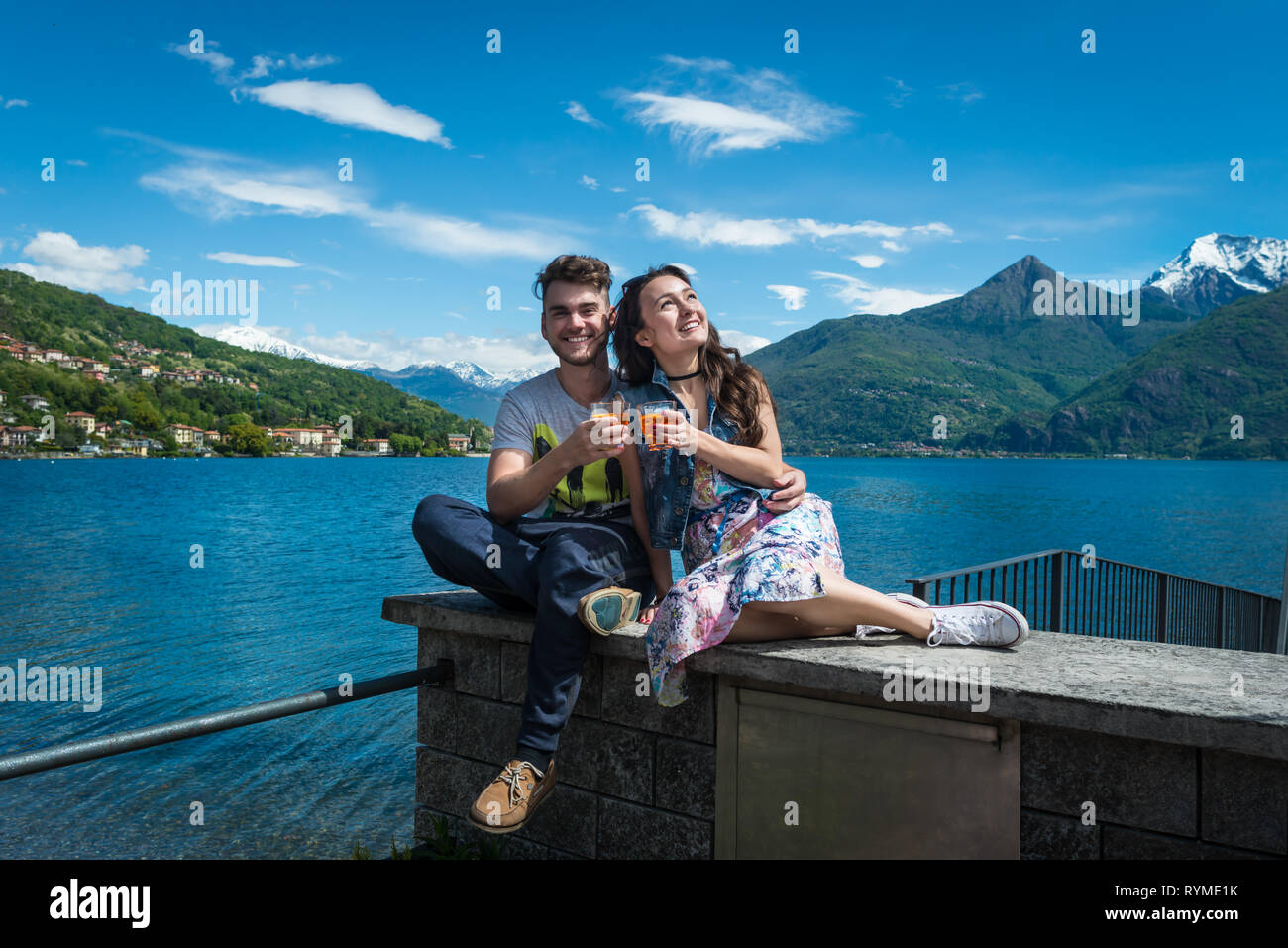 Happy couple sits with two glasses of aperol spritz with scenic view on lake, hills and snowy mountains in Como, Italy. Smiling guy with girlfriend. Stock Photo
