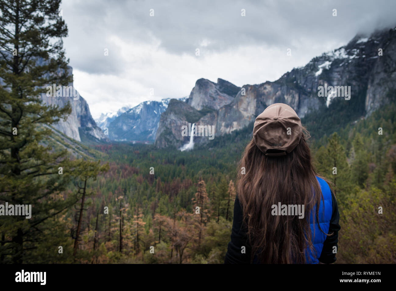 Young female traveler standing back and looking on forest, mountains, waterfall and trees. Girl on background of amazing valley in Yosemite park, USA. Stock Photo