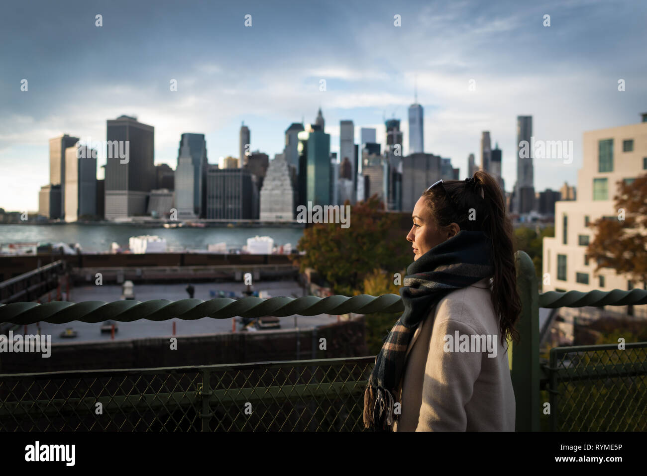 Young woman standing on riverside in Brooklyn and looking on Manhattan skyscrapers. Elegant girl on background of city downtown in New York, USA. Stock Photo