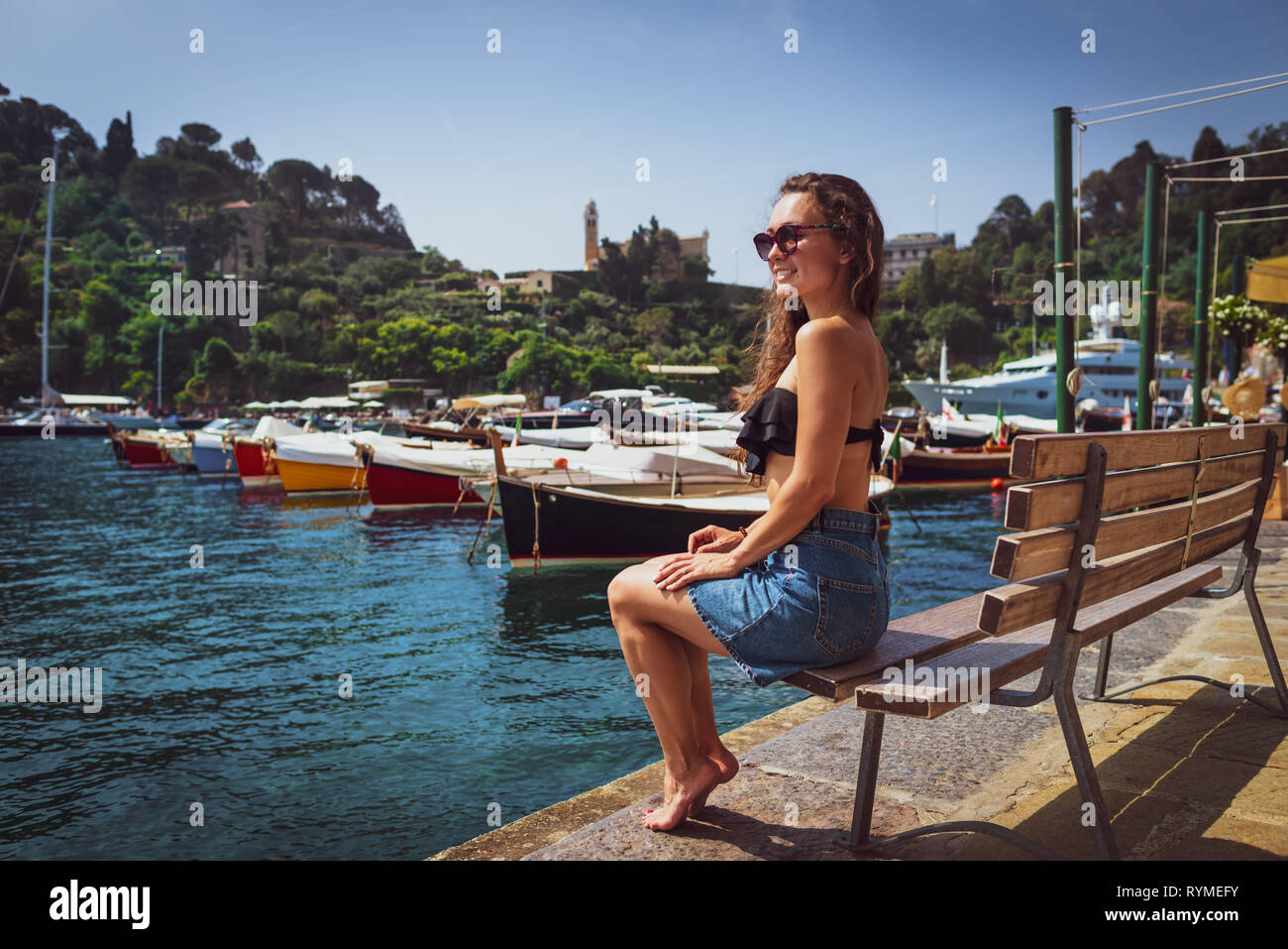 Young barefoot woman sitting on bench in marina in Portofino, Italy. Female tourist enjoying sunny day on background of small village, sea and boats. Stock Photo