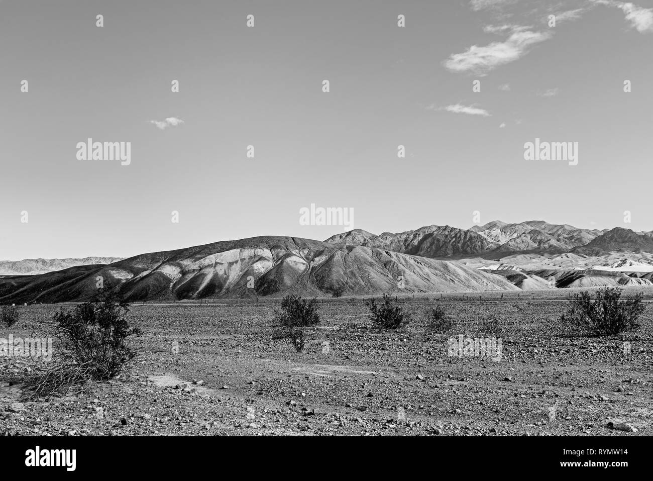 Black and white, sparse vegetation in rocky desert valley with barren hills beyond. Stock Photo