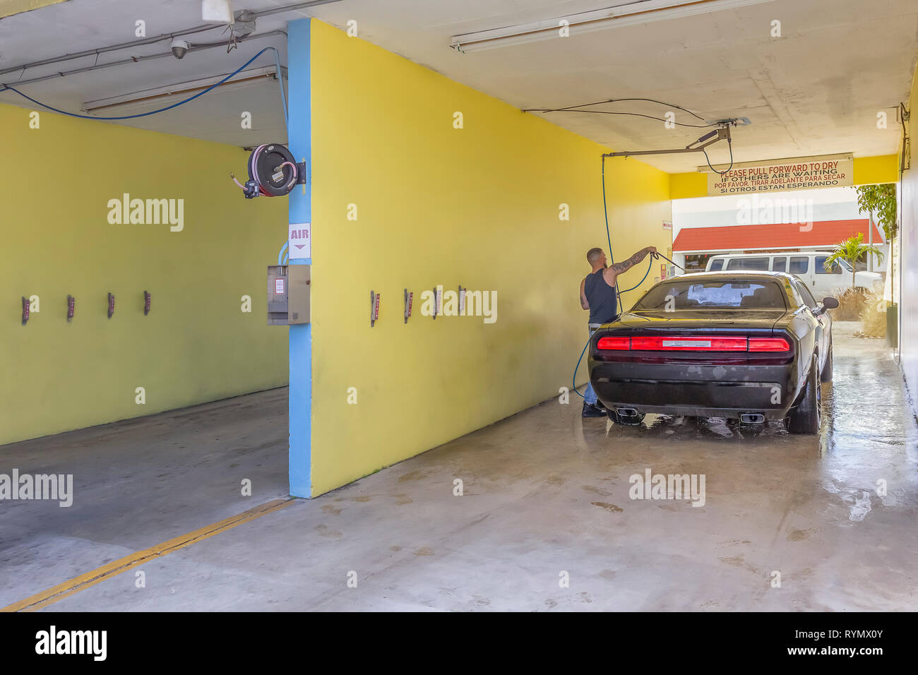 A worker rinses a black car at the car wash. The car parked inside the right bay is being clean by a young man with tattoos. Stock Photo