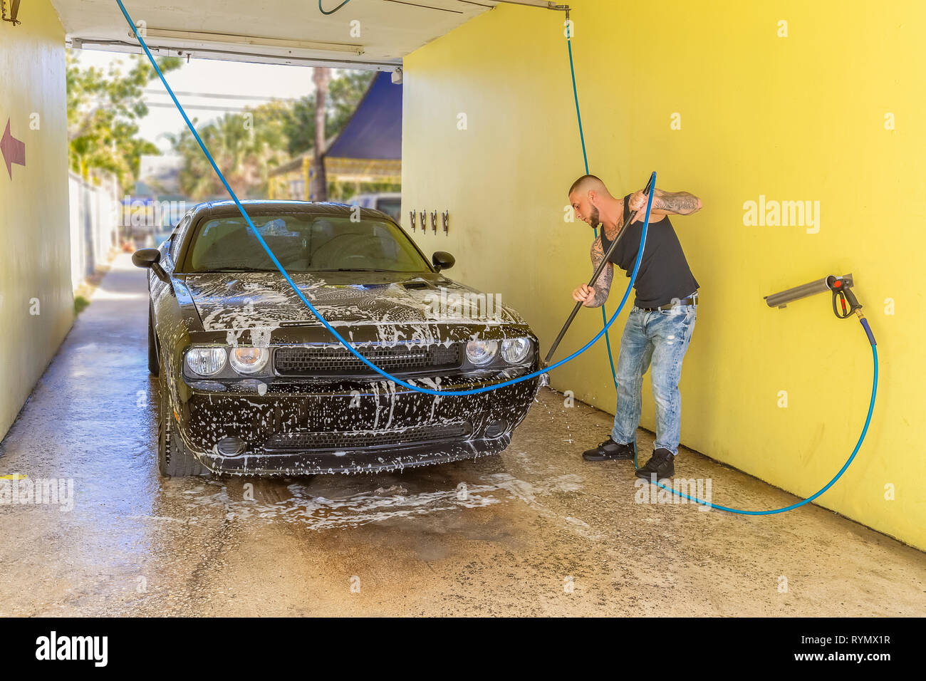 A man is washing his car in carwash bay. The masculine young adult with tattoos soaps up the car by scrubbing every detail. Stock Photo