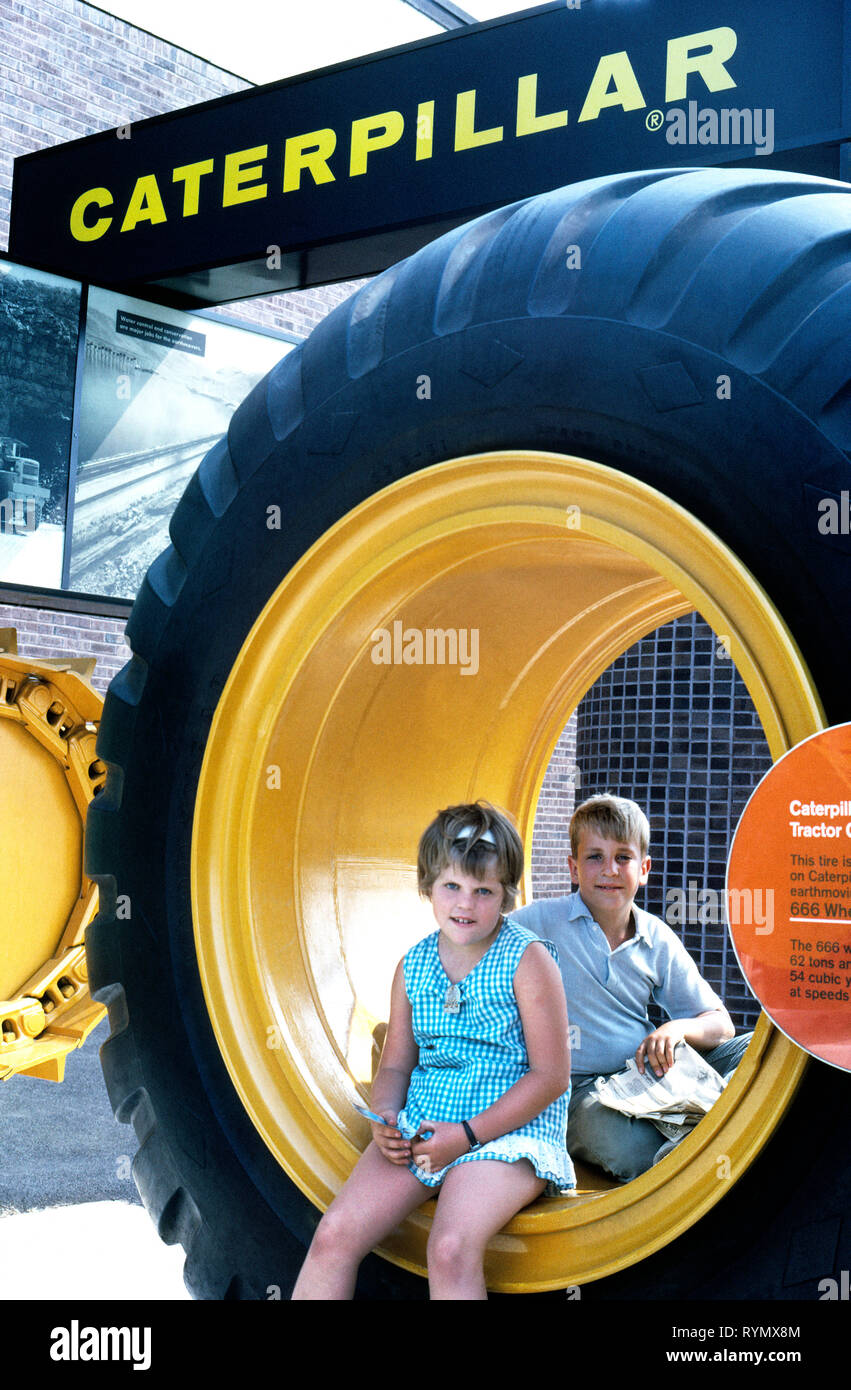 Two young fairgoers rest inside the hub of a huge 10-foot-tall (3-meter) rubber tractor tire in front of the Caterpillar exhibit at the 1964-65 New York World's Fair held at Flushing Meadows Park in the Borough of Queens, New York, USA. Cat is the world’s leading manufacturer of construction and mining equipment, and nearly 200 of its earthmoving machines were used in construction of the 646-acre (261-hectare) fairgrounds. American corporations rather than countries from around the world were the major exhibitors at this largest of the world expositions ever held in the United States. Stock Photo