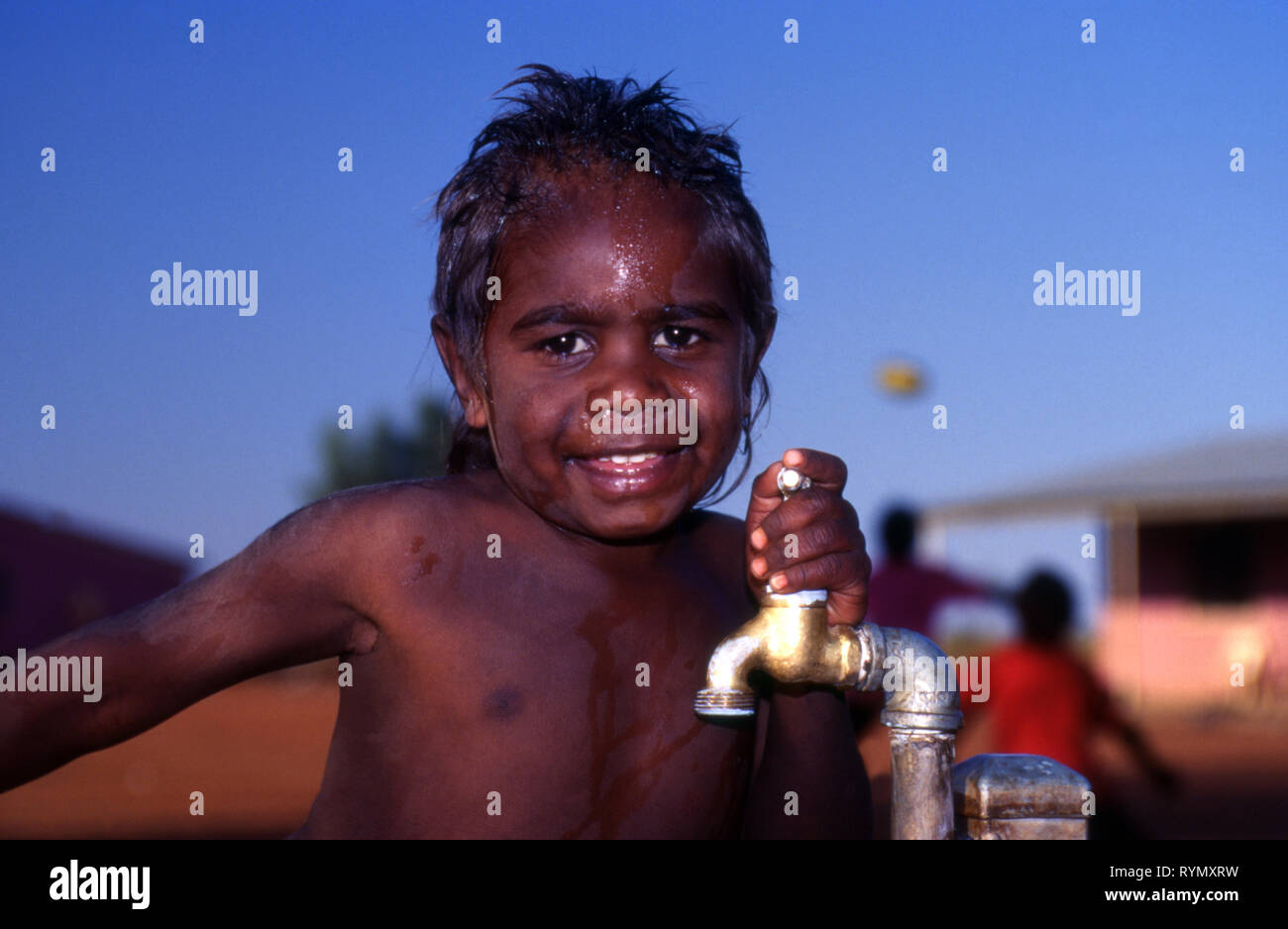 YOUNG SMILING ABORIGINAL BOY HOLDING ON TO A TAP, YUELAMU ABORIGINAL COMMUNITY (MOUNT ALLAN SCHOOL) NORTHERN TERRITORY, AUSTRALIA. Stock Photo