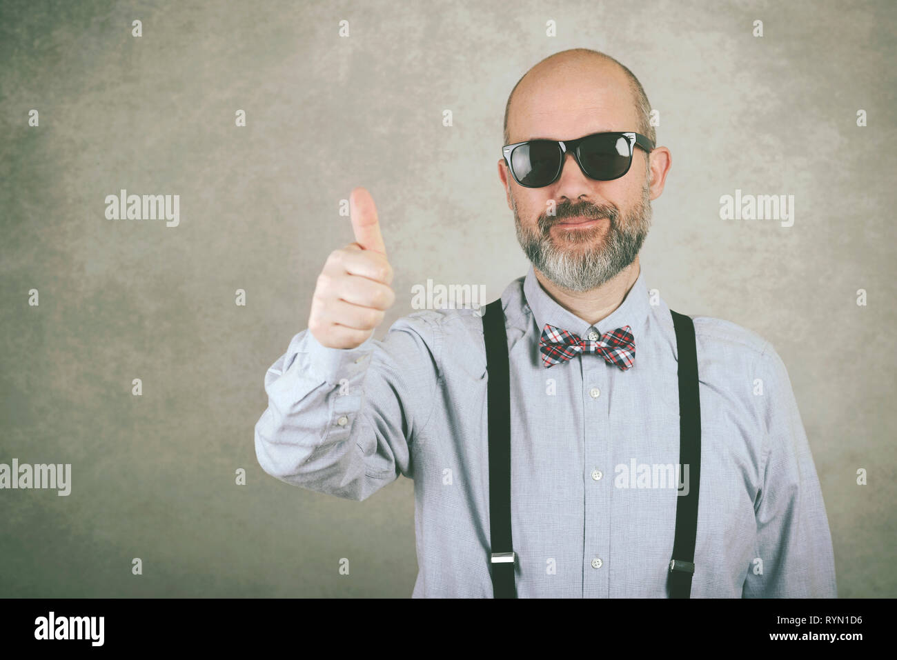 happy man with sunglasses and bow tie showing thumbs up against gray