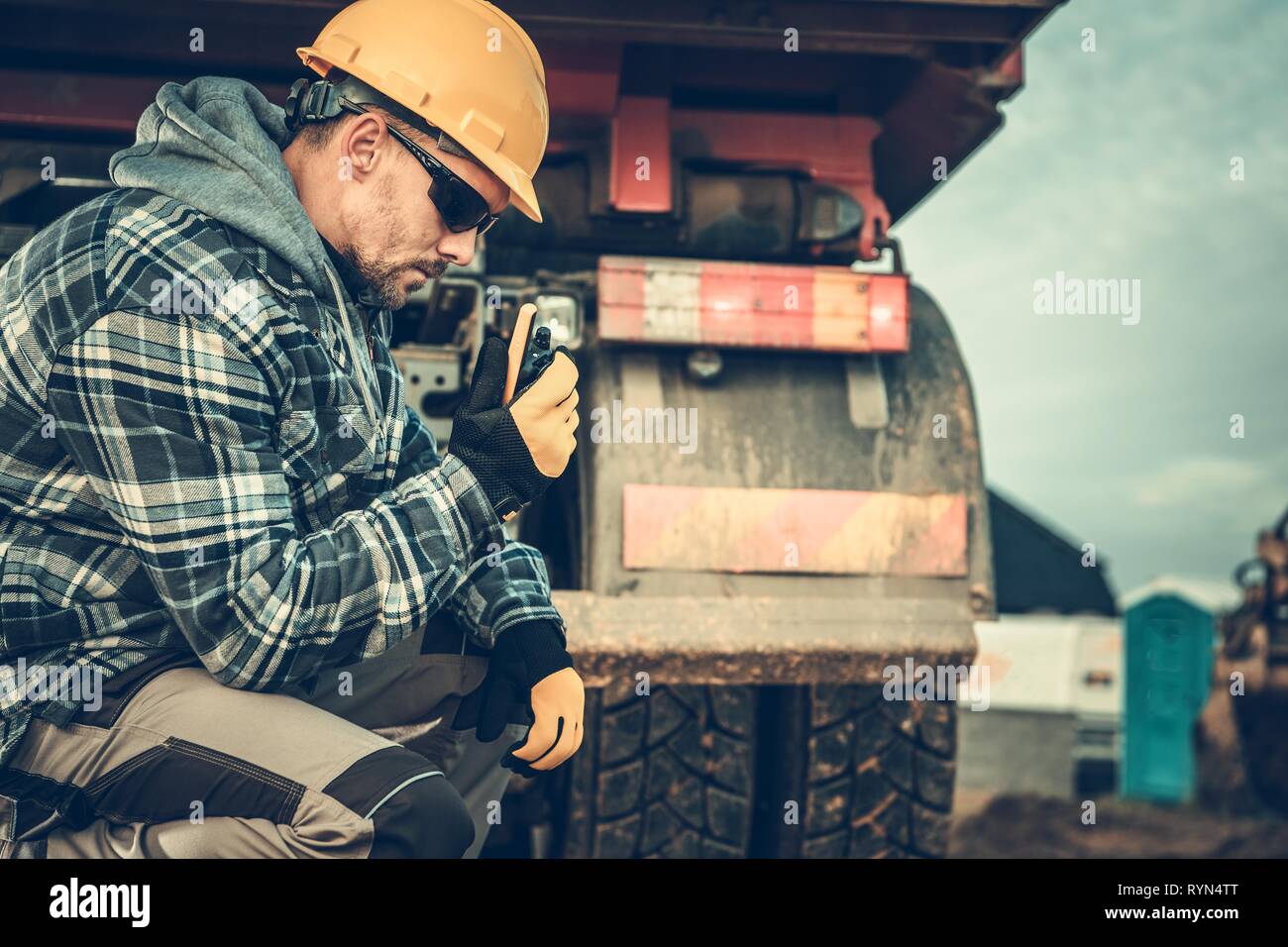 Caucasian Construction Site Worker in Hard Hat and Sunglasses with Walkie Talkie Taking Conversation with Other Building Crew Member. Dump Truck in th Stock Photo