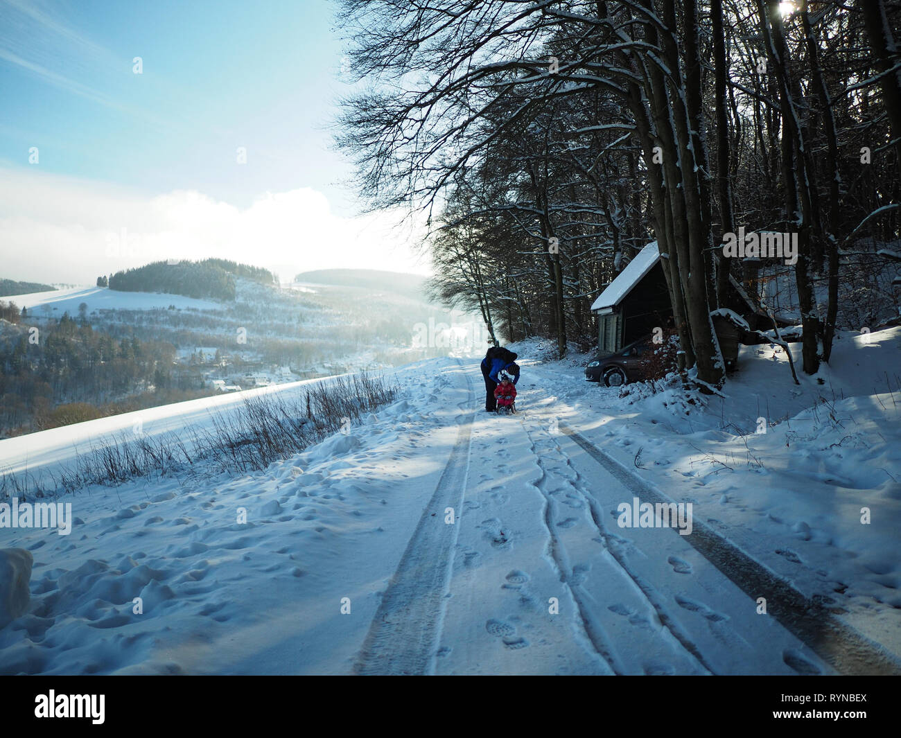 Mother and child with sled on a single track road leading through a snow-covered rural landscape Stock Photo