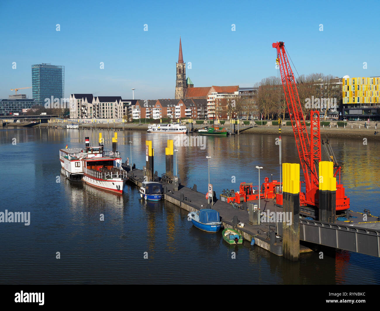Bremen, Germany - February 14th, 2019 - Pier with several small vessels and bright red floating crane and city skyline with Weser Tower and St, Stepha Stock Photo