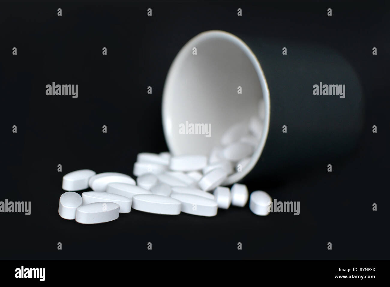 Many white medical pills spilling from a black bottle container on dark black background Stock Photo