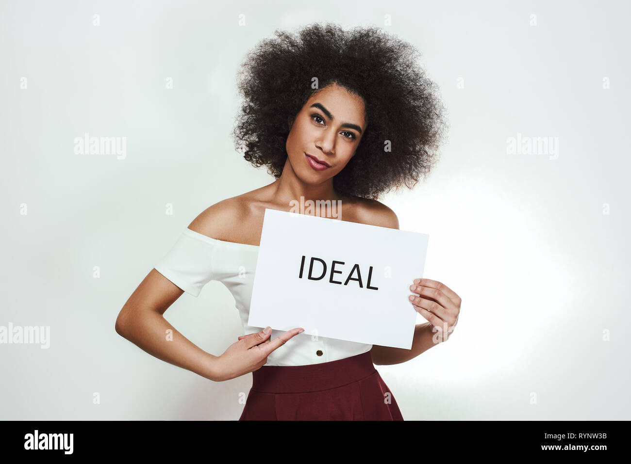 Perfect in everything. Studio portrait of confident young afro american woman holding a paper which is saying 'Ideal' while standing against grey background. Studio portrait Stock Photo