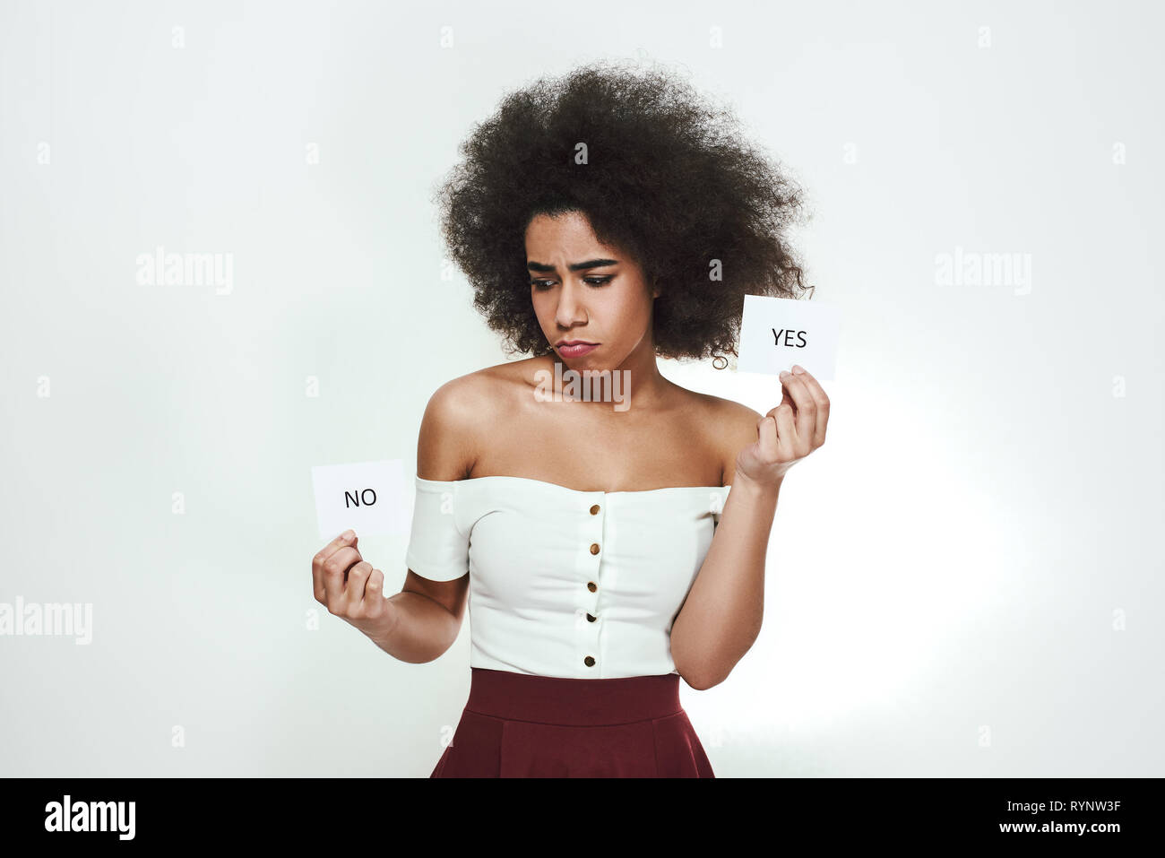 I am not sure...Confused young afro american woman is holding papers with yes and no on them and thinking while standing against grey background. Doubt concept. Thinking process Stock Photo