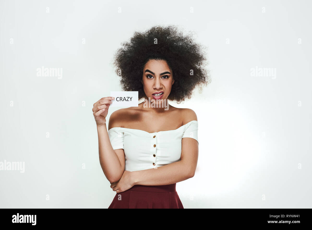 Going crazy! Young angry afro american woman making a mad face and holding a piece of paper which is saying 'crazy' . Studio shot. Moody. Text message Stock Photo