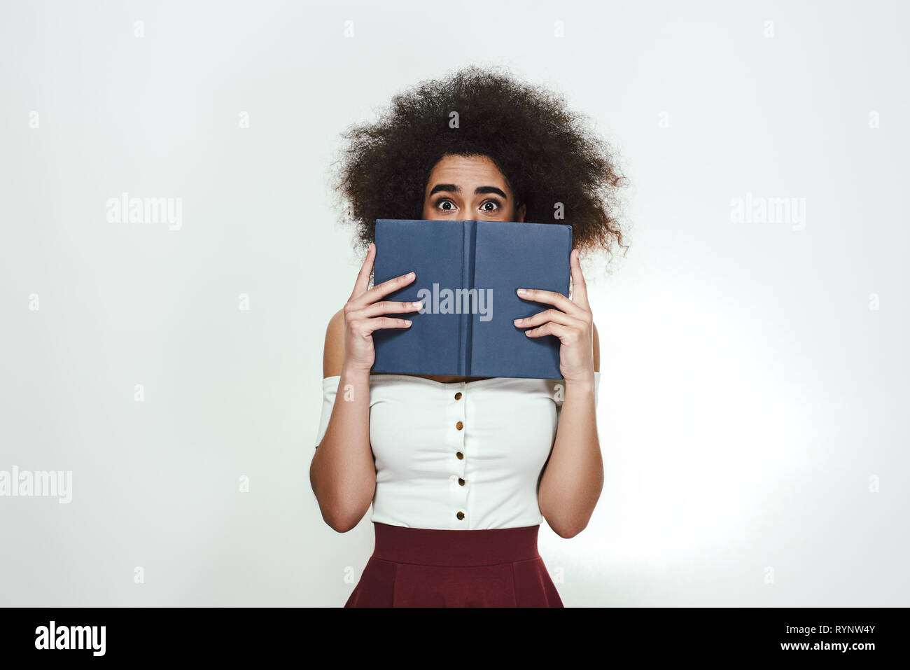 Worried about her exams. Frustrated young african woman holding book and covering her face with it while standing against blackboard. Book reading. Learning concept Stock Photo