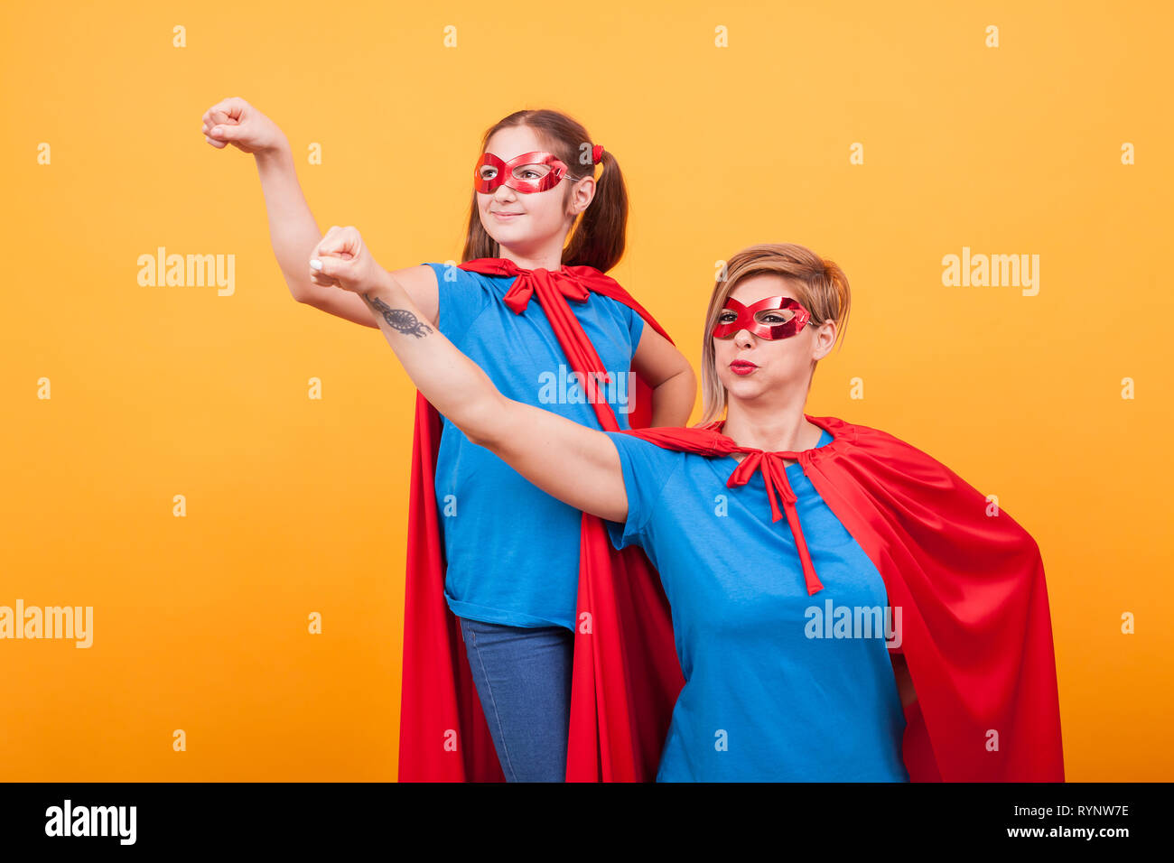 Mother and daughter dressed like heroins holding their fist in the air and looking away over yellow background. Happy childhood. Mom super powers. Stock Photo