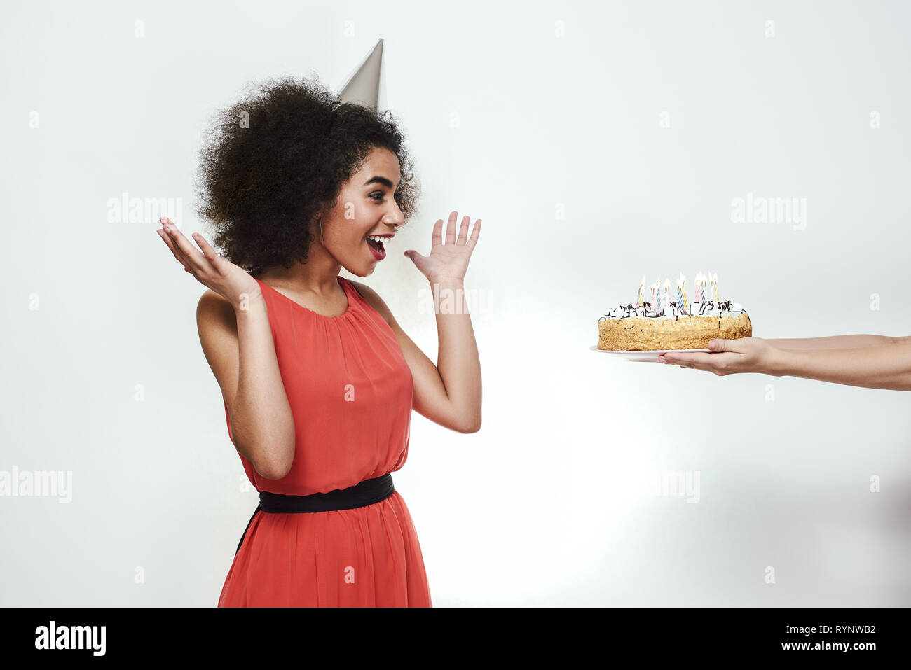 Surprise for you! Excited young afro american woman in party hat raising up her hands and looking at birthday cake with candles Stock Photo