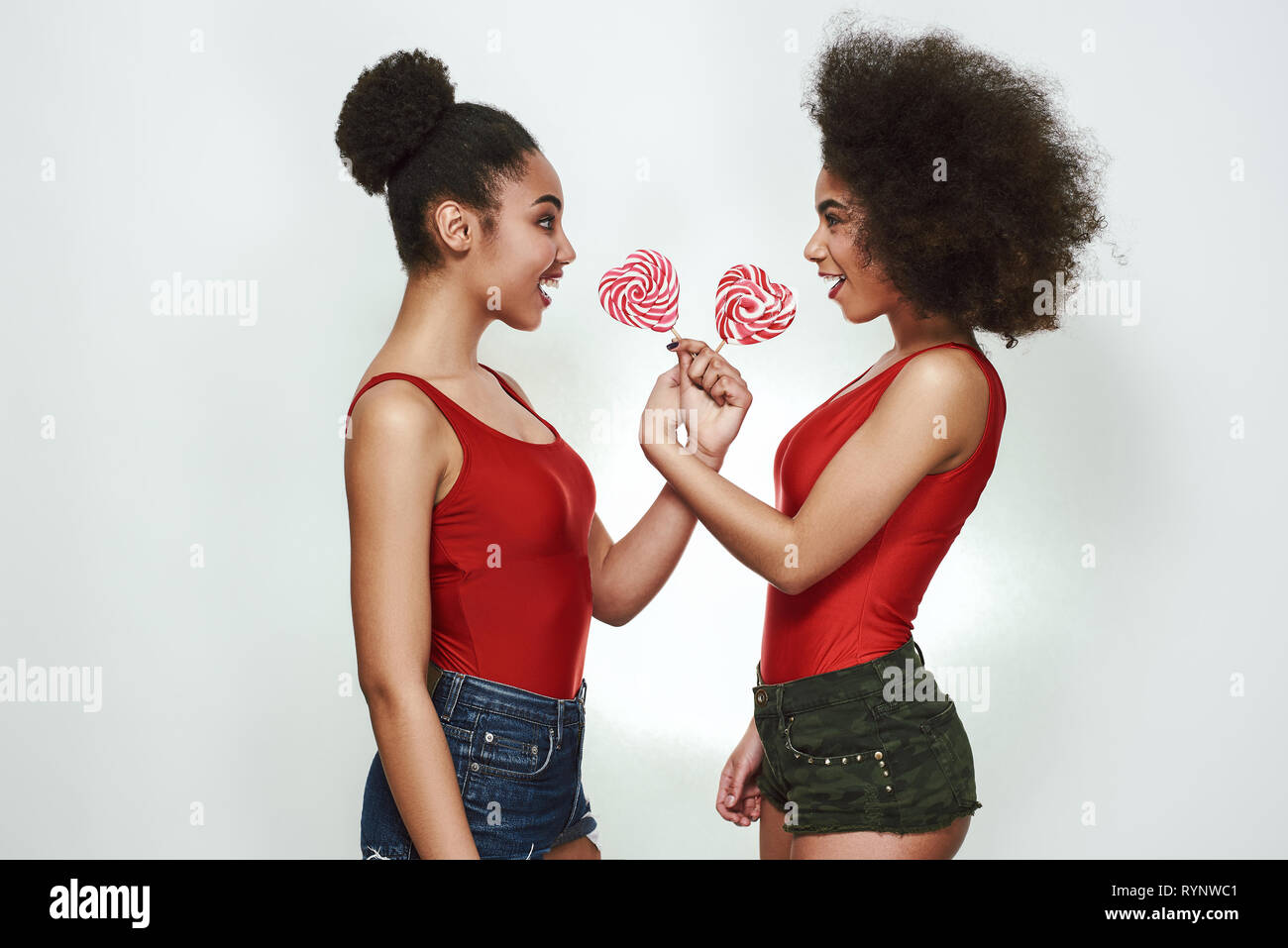 Try my lollipop! Two cheerful young afro american woman in summer clothes are holding lollipops and looking at each other while standing against grey background. Fun concept. Women beauty Stock Photo