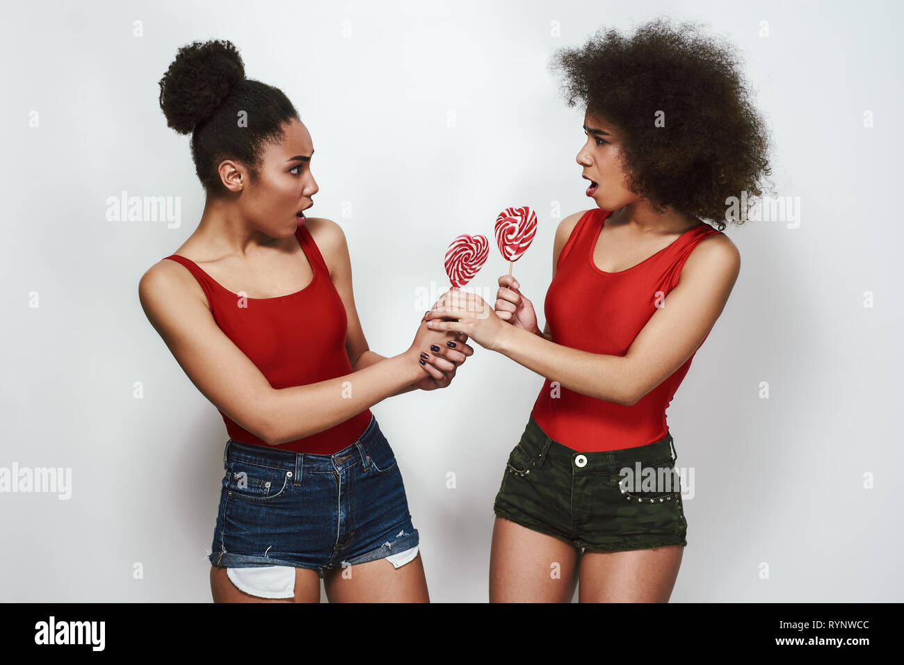 I want more! Two angry afro american woman in jeans shorts are looking at each other with opened mouth and holding lollipops shaped heart while standing against grey background Stock Photo