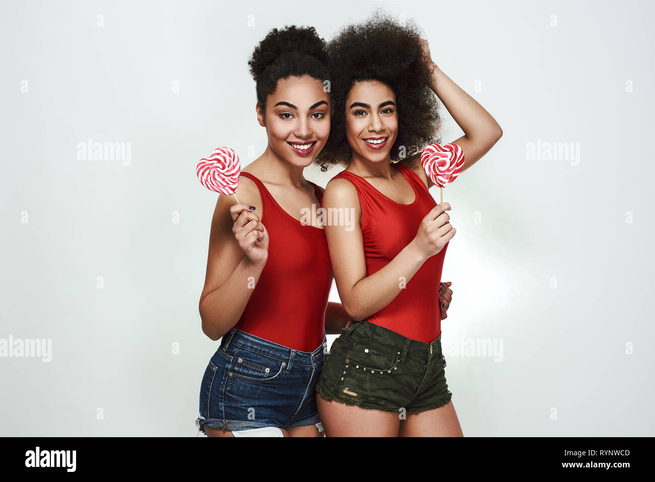 Sweet beauty. Two beautiful young afro american women in summer clothes holding lollipops shaped heart and smiling while standing against grey background Stock Photo