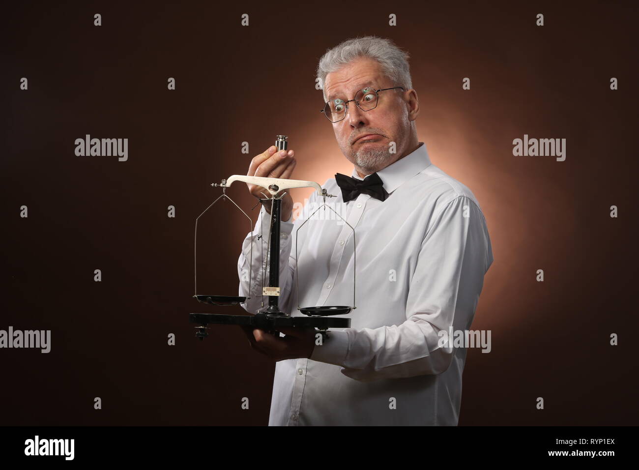 Elderly gray-haired man 50s, in white shirt, glasses and bow tie weighing something on scales with small kettlebells Stock Photo
