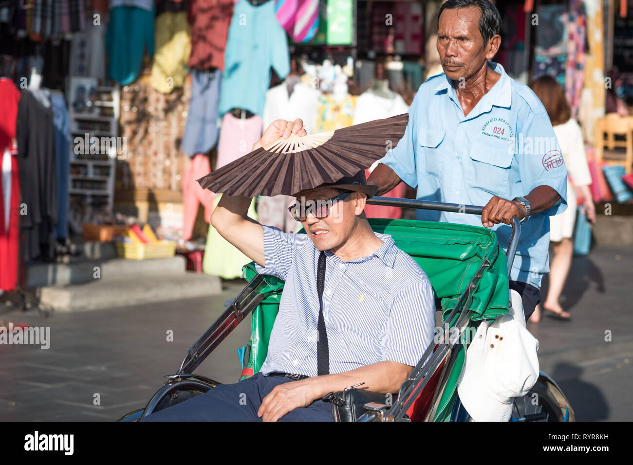 Hoi An, Vietnam - October 23, 2018: an Asian man covers his face with a fan trying to defend his eyes against harsh sunlight at a rickshaw's cabin. Stock Photo