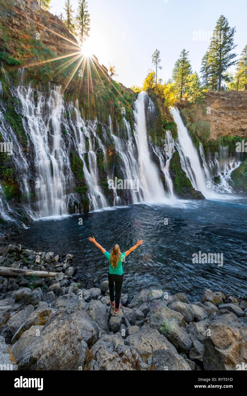 Young woman standing at a waterfall, stretching arms into the air, McArthur-Burney Falls Memorial State Park, California, USA Stock Photo