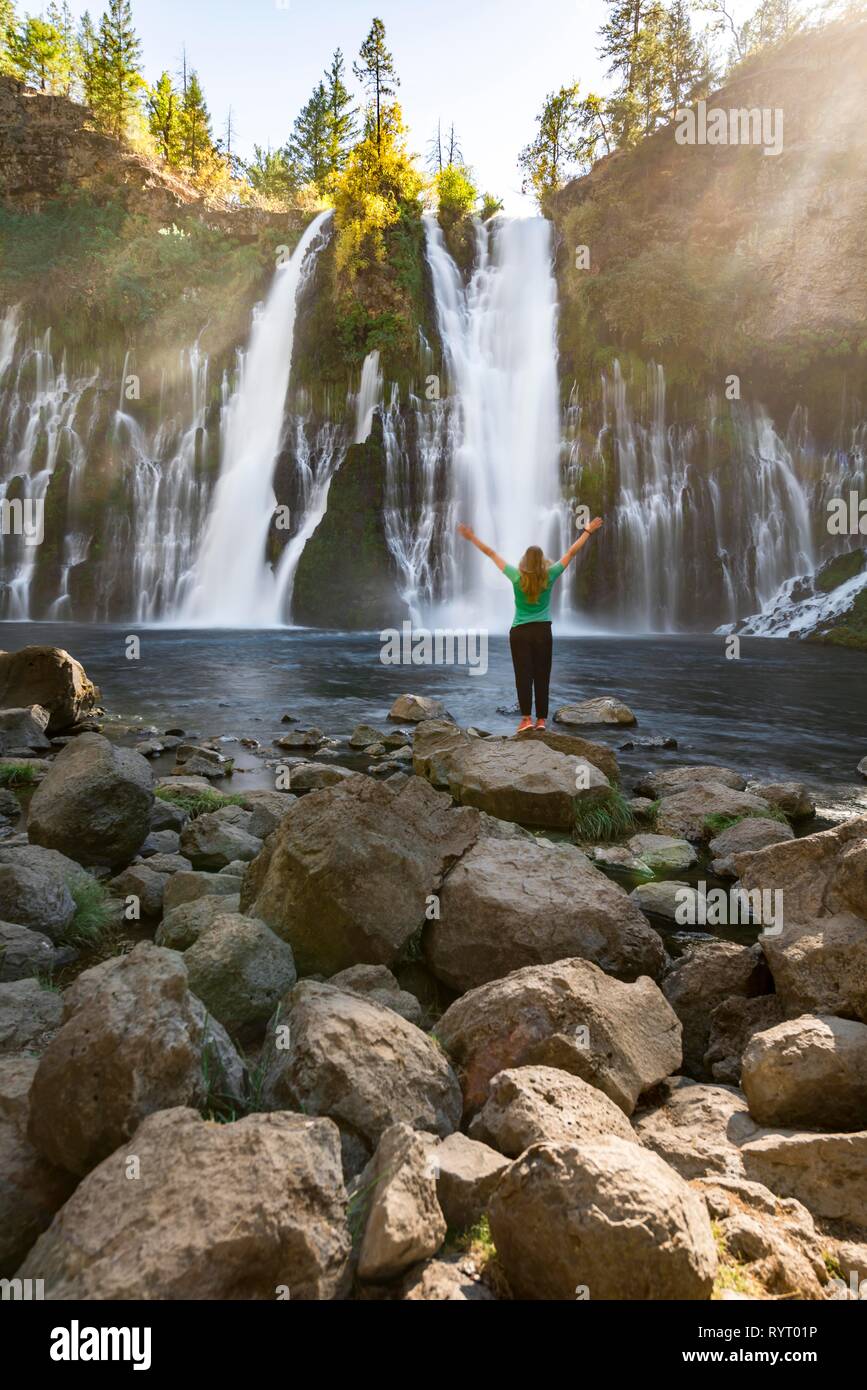 Young woman standing at a waterfall, stretching arms into the air, McArthur-Burney Falls Memorial State Park, California, USA Stock Photo