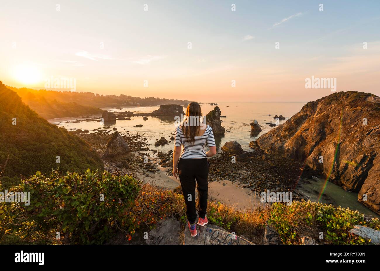Young woman looking into the sunrise, rugged coastal landscape with many rocky islands, Arch Rock, Harris Beach State Park Stock Photo