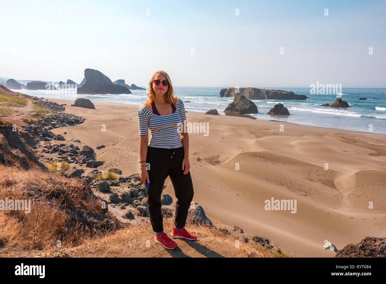 Young woman looking into the camera, sandy beach and coast with rugged rocks, Myers Creek Beach Viewpoint, Oregon USA Stock Photo