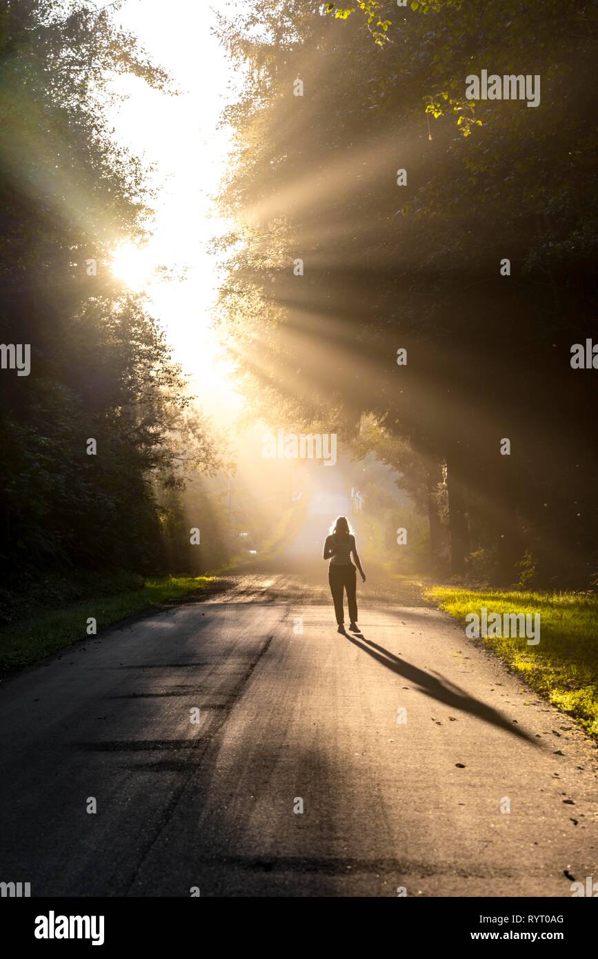 Young woman walking on a street, sunlight shining through trees, Oregon Coast Highway, Oregon, USA Stock Photo
