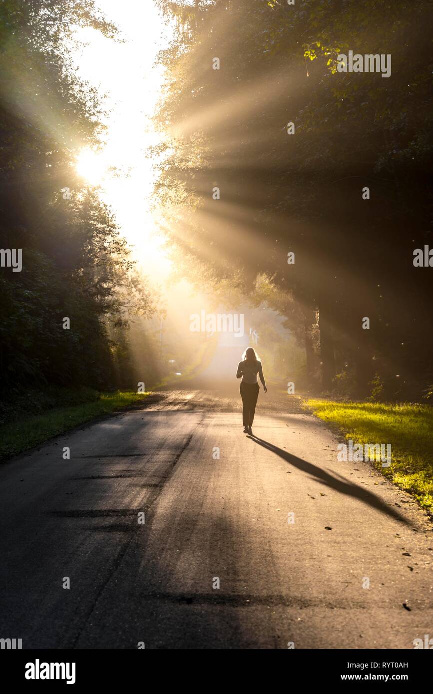 Young woman walking on a street, sunlight shining through trees, Oregon Coast Highway, Oregon, USA Stock Photo